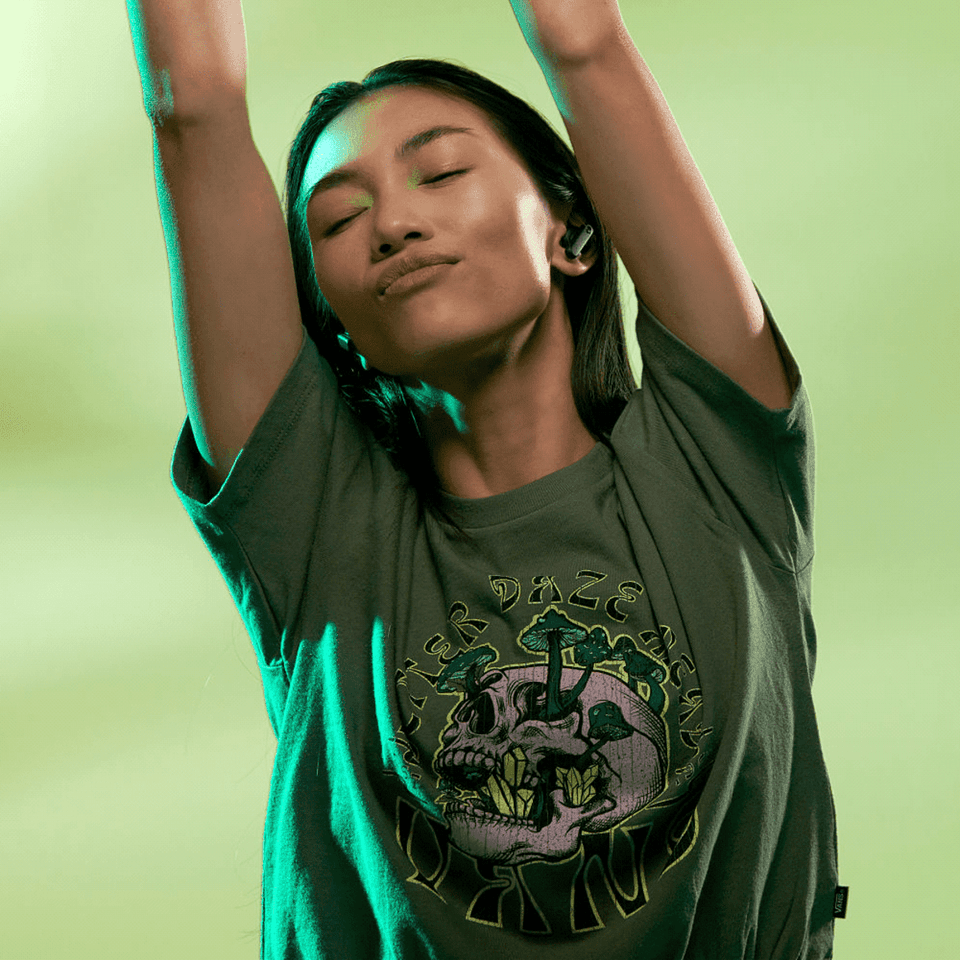 Earbuds in dark colour being worn by a model in a graphic tee, with a vibrant green background.