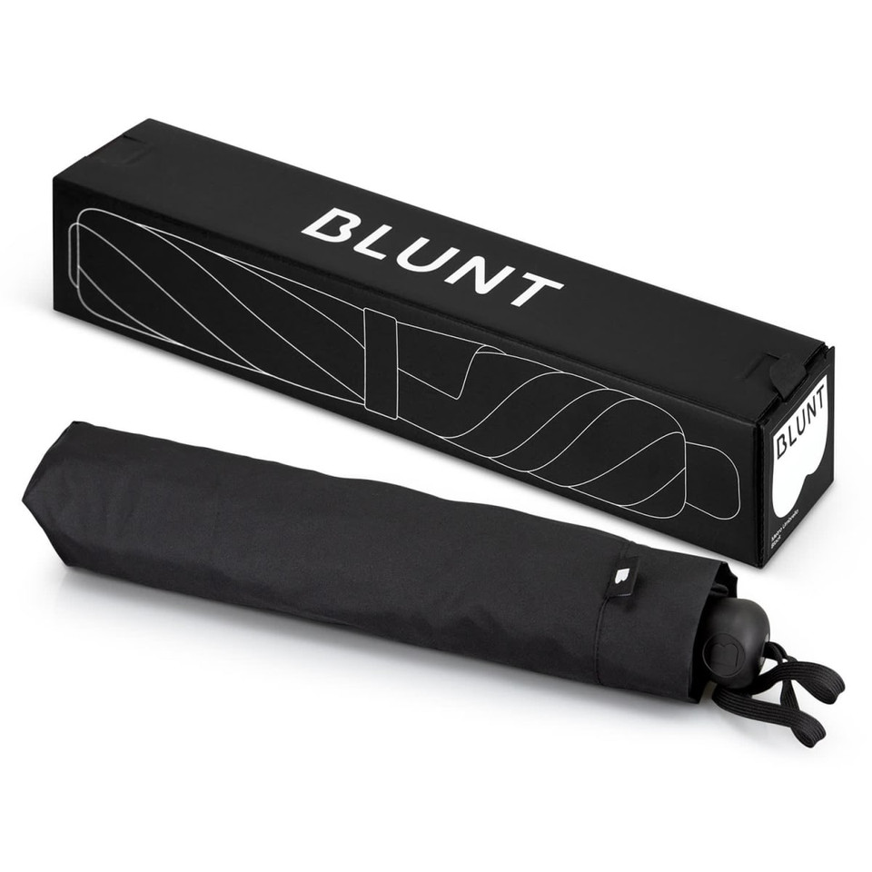 A black BLUNT Metro Umbrella laid flat next to its rectangular packaging, featuring a logo.
