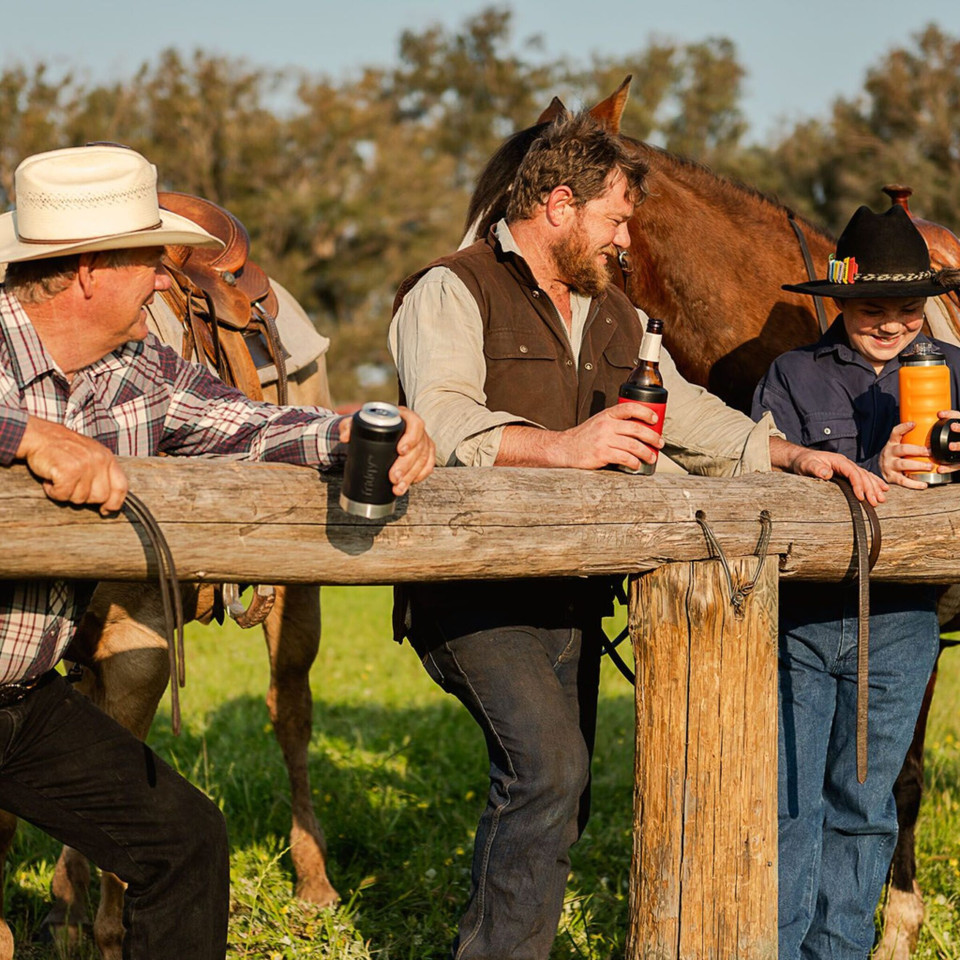 Three people standing behind a wooden fence. An elderly gentleman, a middle aged bearded man and a young lady with black widebrimmed hat. Behinde the people are two horses partially visible. The two gentlemen are holding fridgy stubby holder. One with a c