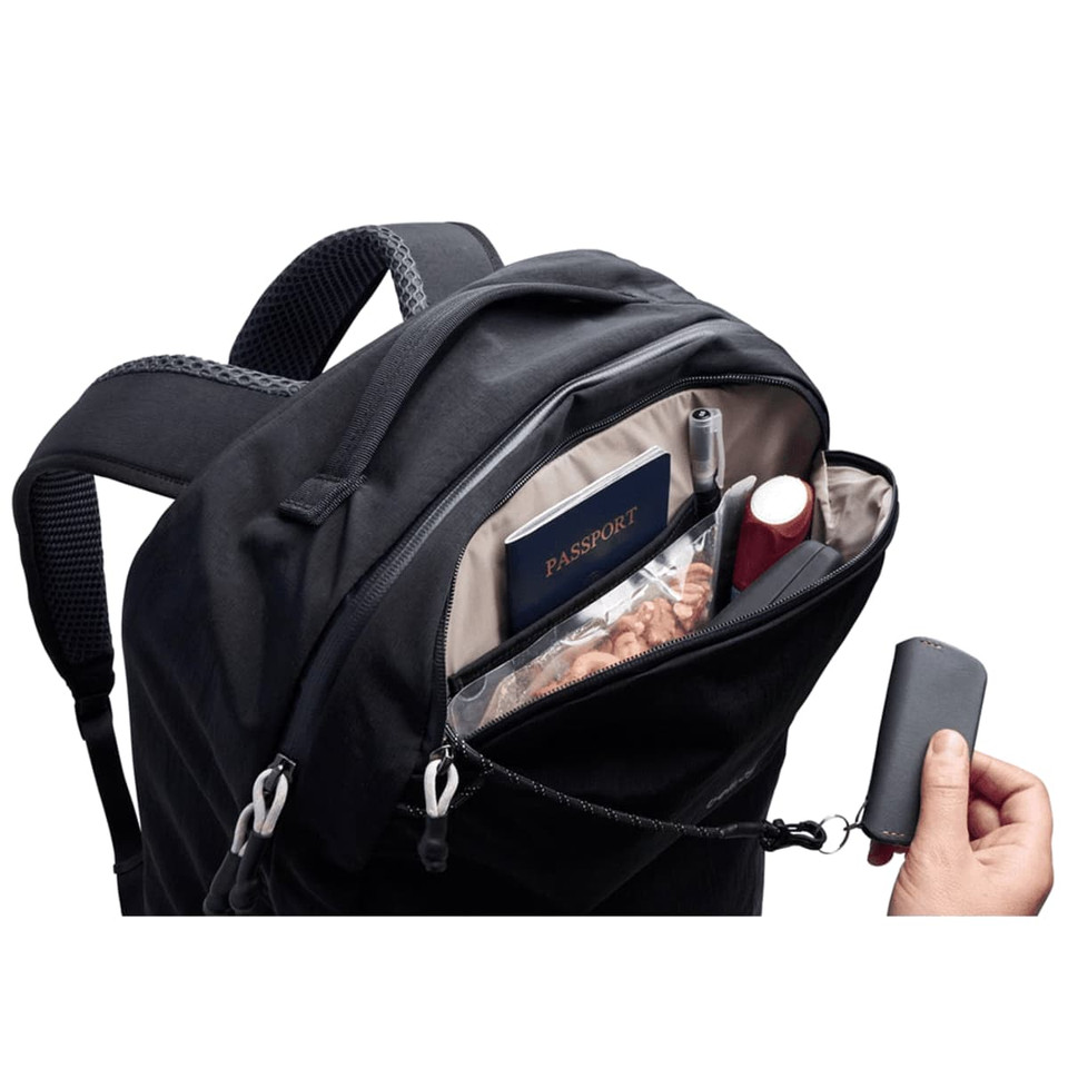 A black backpack with a zipped pocket containing a passport, pens, and a small bottle. It has a branding logo.