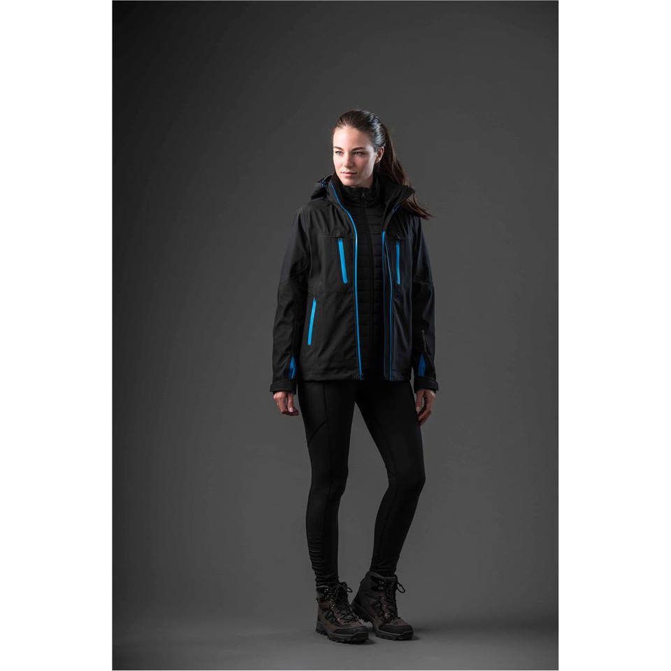 A woman in a black jacket with blue accents stands against a dark background, wearing black leggings and boots.