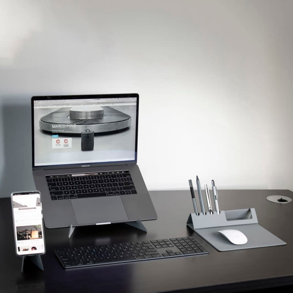 A sleek grey Ascend Mini Stand holding a smartphone, positioned beside a laptop, keyboard, and pen holder on a desk.
