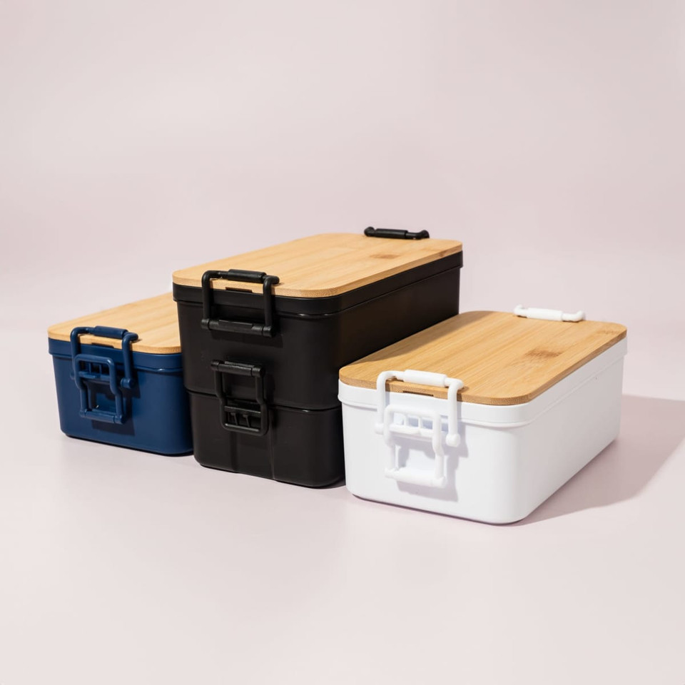 Three lunch boxes in navy blue, black, and white with bamboo lids and handles, displaying a sleek, modern design.