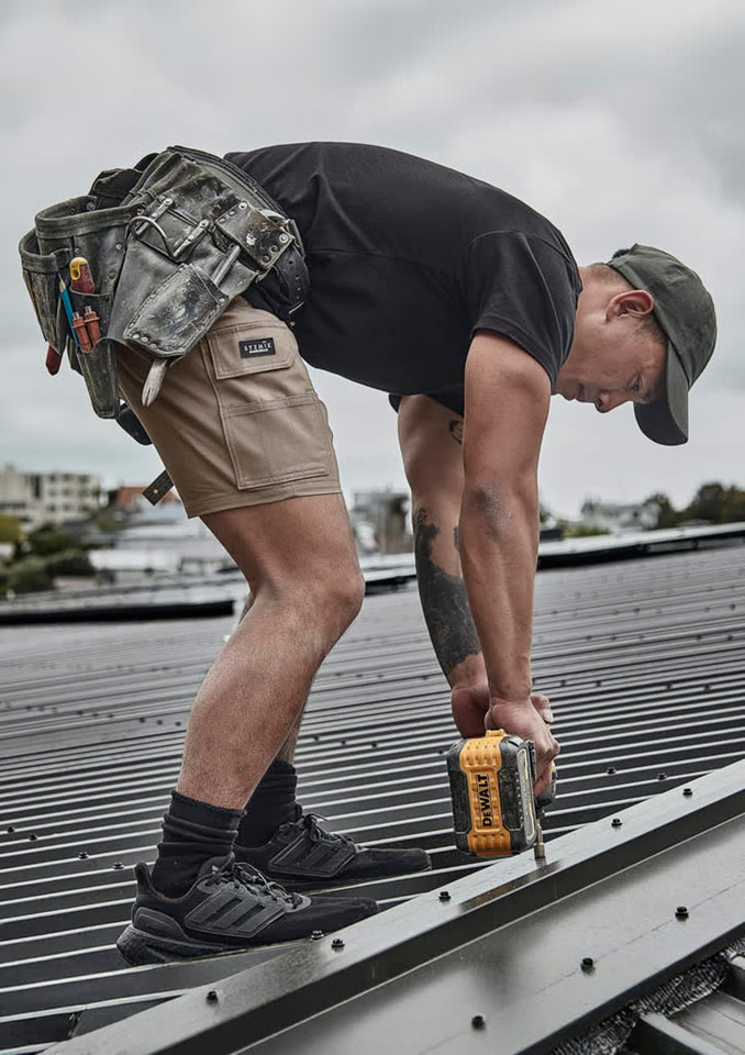 A person wearing khaki cargo shorts and a black t-shirt is using a power drill on a roof. Tools hang from a tool belt.