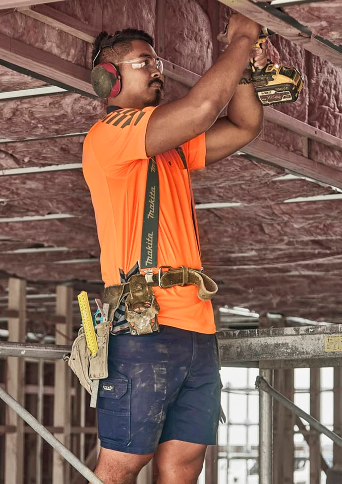 A man wearing an orange shirt and navy blue cargo shorts works on a construction site, using a power drill.