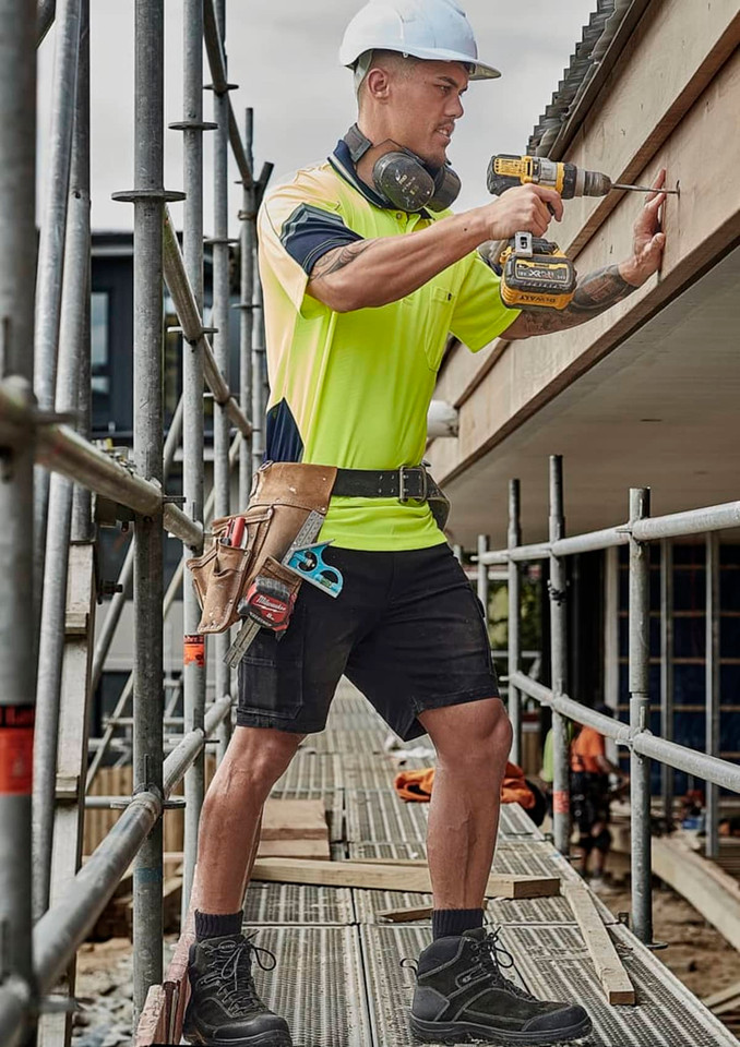 A man in a high-visibility shirt and cargo shorts uses a drill on a construction site, wearing safety gear and a tool belt.