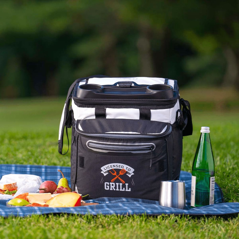 A black and white rolling cooler with a logo, placed on grass near picnic food and drinks.