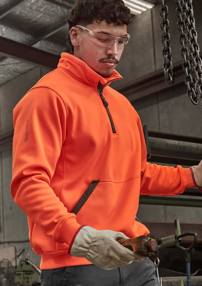 An orange unisex 1/4 zip pullover is worn by a person inside a workshop, featuring protective glasses and gloves.