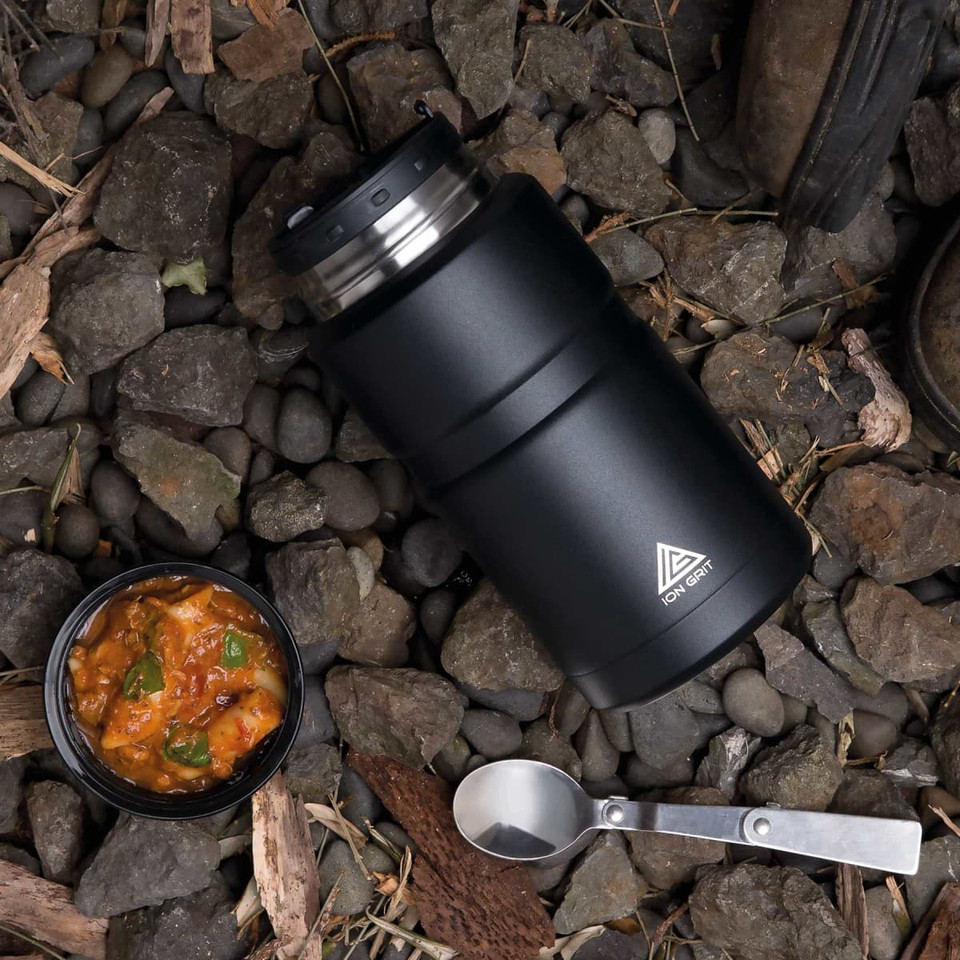A black food jar sits on rocky ground, with a bowl of stew and a spoon nearby. The jar features a logo.