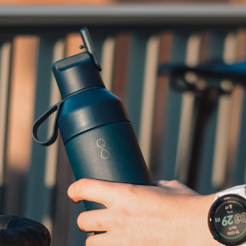 A drink bottle in dark blue, with a handle and a logo, held in a hand near a bike.