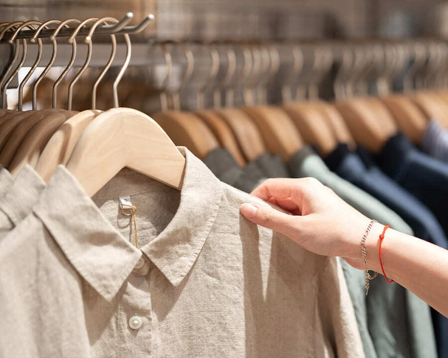 A hand browses beige shirts on wooden hangers in a clothing store