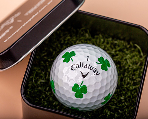A callaway branded golf ball with green shamrocks sits in a black box on a bed of green moss