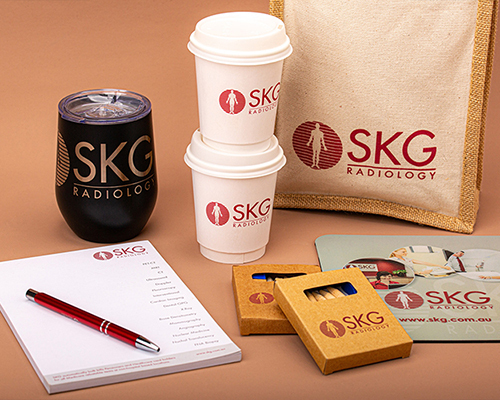 A collection of skg radiology-branded items on a table: two stacked coffee cups, a black tumbler, a notebook, pen, matchboxes, and a tote bag