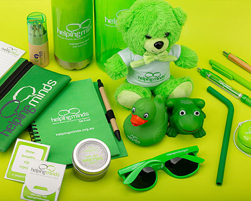 Bright green helping minds-branded promotional items on a yellow background, including a teddy bear, rubber duck, notebook, pen, tin, sunglasses, and water bottle