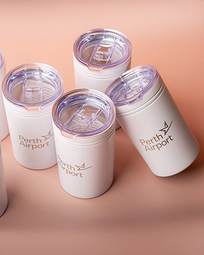 Five white perth airport-branded travel mugs with clear lids are arranged on a soft pink surface