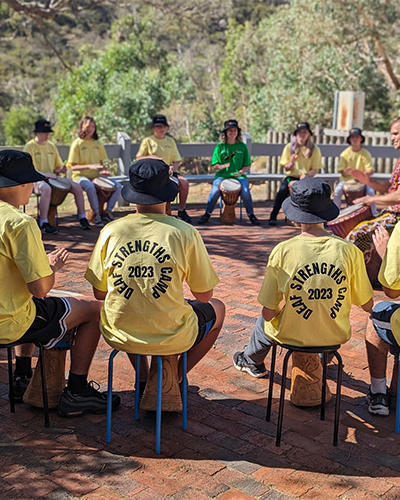A diverse group of people sits in a circle outdoors, each wearing a yellow "deaf strengths camp 2023" t-shirt playing drums