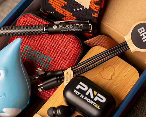 A group of branded promotional products in a packaging box, inc a custom stress ball, charging cable, phone stand, speaker and ear bud