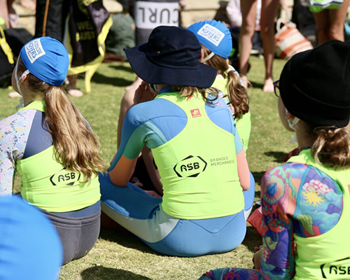 A group of young girls in yellow and blue wetsuits sitting on the grass