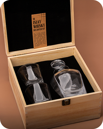 A wooden box with an open lid contains an elegant glass whisky decanter and two glasses, set against a dark lining