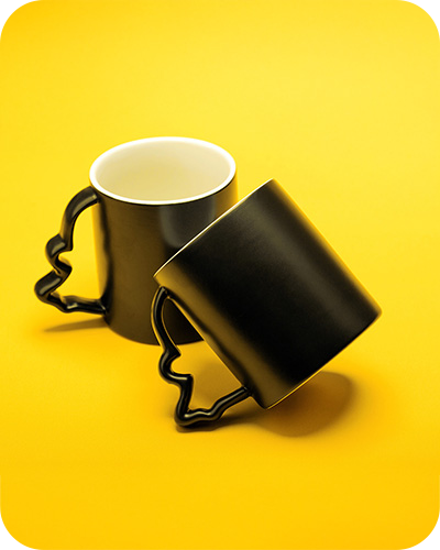 Two black coffee mugs with silhouette handles of a face against a vibrant yellow background