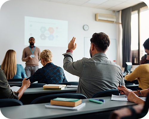 A classroom setting with a man standing in front of a presentation and a person seated raises a hand