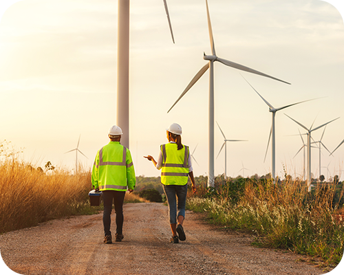 Two people in safety gear and helmets walk on a dirt path towards towering wind turbines