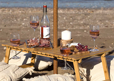 A beach picnic setup with a wooden table holding a wine bottle, glasses of rosé, a candle, grapes, and charcuterie