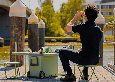 A person sits by a river, drinking from a can next to a small portable cooler with bottles and snacks