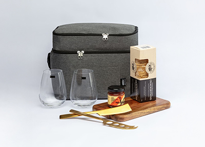 A grey insulated picnic bag, stemless wine glasses, a jar, gourmet crackers, and a cheese knife on a wooden board