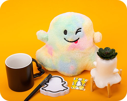 A collection of snapchat branded merchandise, including a pastel tie-dye plush toy  surrounded by a black mug, a pen, notepad, cards, and more