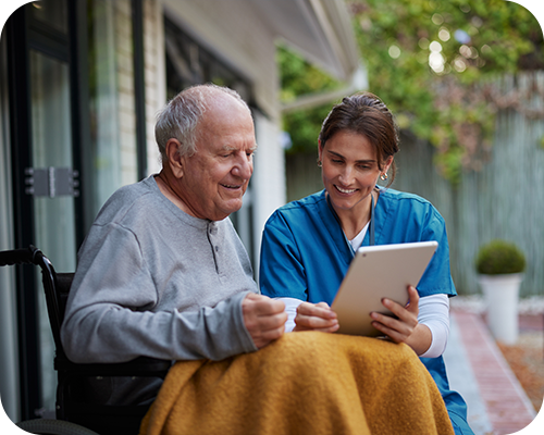 Elderly man in a wheelchair, covered with a yellow blanket, smiles while a nurse in blue scrubs shows him a tablet