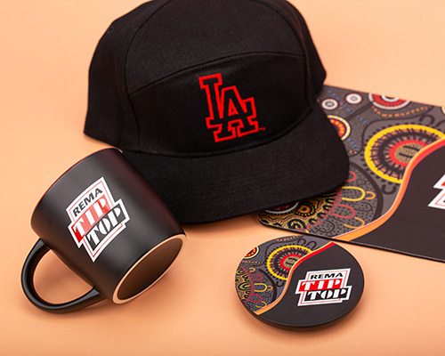 Black cap, black mug with rema tip top branding, and a coaster on a colourful, patterned surface