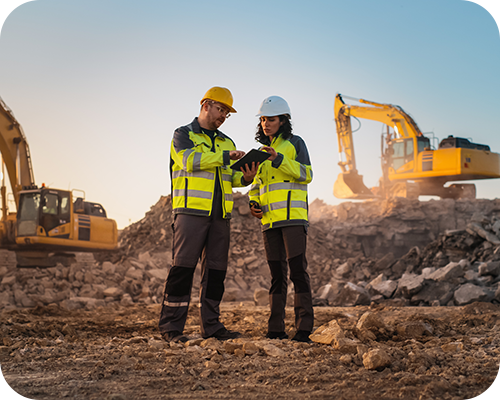 Two construction workers in safety gear discuss plans at a rocky site