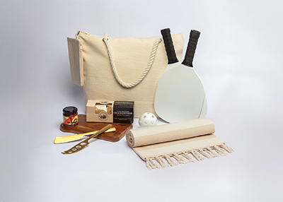 A beige tote bag, paddles, a ball, rolled towel, cheese knife, gourmet items, and wooden tray
