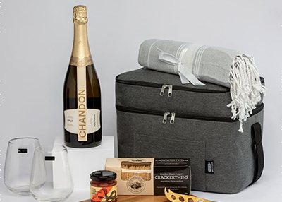 Picnic set with a champagne bottle, stemless glasses, a grey cooler bag, a blanket, crackers, and a jar