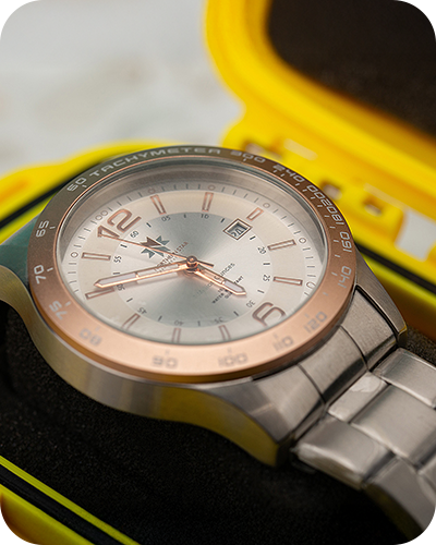 A sleek silver watch with a copper-toned bezel and white face sits in a vibrant yellow case