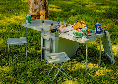 A portable picnic table set on grass, with folding stools, canned drinks, bags of chips, and an ice bucket