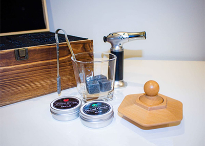 A cocktail smoking kit with a wooden lid, torch, and metal spoon on a white table