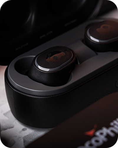 A close-up of black skullcandy wireless earbuds in an open charging case. Surrounding are black cases labeled with conocophillips