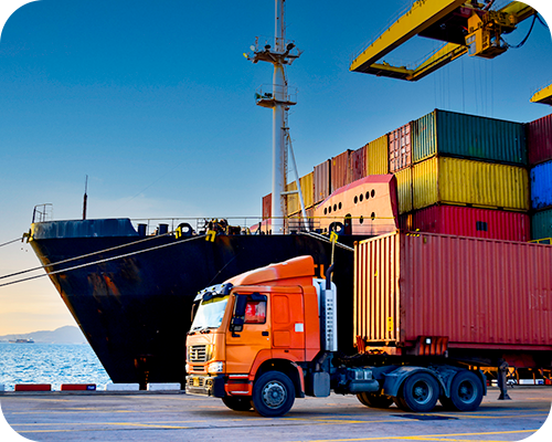 Cargo ship docked at port with stacked containers, a large crane, and an orange truck loaded with a container