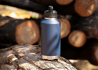 A blue insulated bottle sits on a log with stacked wooden logs in the background