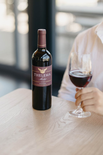 Bottle of Thelema Merlot, Stellenbosch 2020 red wine with plum and spice flavors. Bottle next to person holding glass of red wine.