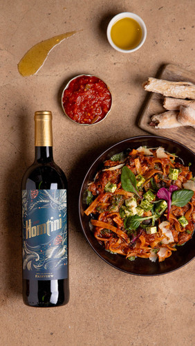 Bottle of Fairview Homtini, Coastal Region 2022 wine with berry and spice flavors. Bottle of wine next to plate of food.