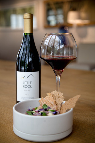 Bottle of Mont Rochelle Little Rock Rouge, Franschhoek 2022 red wine with dark fruit and spice. Bottle of wine next to glass of red wine and plate of food.