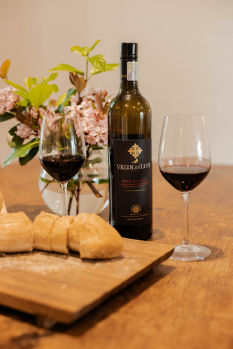 Bottle of Vrede en Lust Boet Erasmus Red Blend, 2019 from Simonsberg-Paarl with notes of Red and black fruit, tobacco and vanilla. Bottle of wine between two glasses of red wine, next to bread rolls on a wood board.