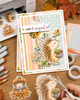 Pre-coloured printable clipart of a cute woodland hedgehog holding a bowl filled with acorns and Fall leaves, surrounded by softly coloured falling Autumn leaves and acorns in a delicate watercolour style.