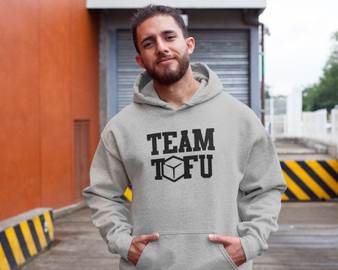 Young man standing at a loading dock, wearing a sport grey unisex vegan hoodie with the words Team Tofu printed on it.