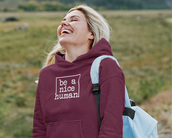 Young blonde woman laughing while carrying a backpack and wearing a maroon hoodie that says be a nice human.