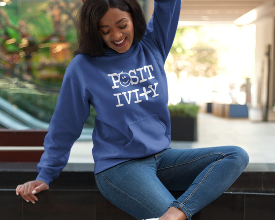 Young smiling woman sitting on a ledge while wearing a royal blue hoodie with the word positivity with a smiley face and plus sign.