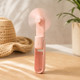 Pink foldable USB rechargeable handheld fan standing upright on a wooden table with summer accessories in the background.